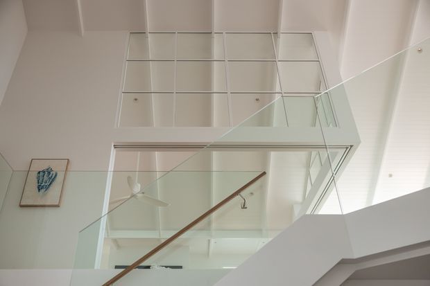 Upward-looking interior view of a bright contemporary stairway or landing, featuring a glass balustrade, timber handrail, white structural elements, and a large overhead mirrored or glazed grid feature. A ceiling fan and framed artwork are visible in the background.