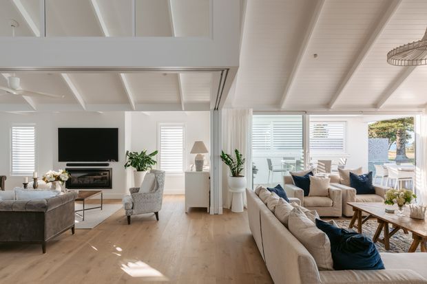 Bright open-plan coastal living room with white-painted exposed ceiling beams, pale timber flooring, large windows with plantation shutters, and a relaxed seating arrangement around a fireplace and wall-mounted television. A dining area is visible beyond the living space, with a neutral palette accented by navy cushions and indoor greenery.