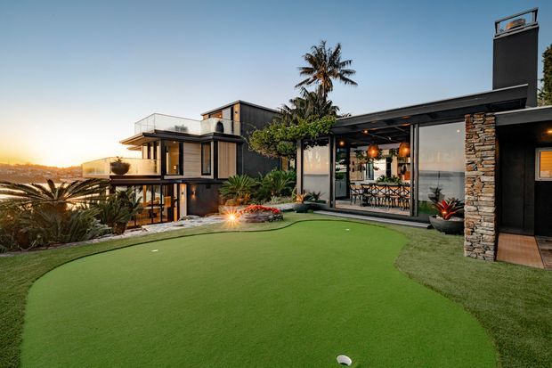A modern multi-level residence photographed at sunset, featuring extensive floor-to-ceiling glazing, dark cladding, projecting balconies, a broad low-pitched roof, and a prominent stone-clad chimney. The landscaped foreground contains a manicured artificial putting green, while a glazed outdoor entertaining area opens toward a distant coastal or urban view. Palm trees and layered planting reinforce the warm outdoor setting.