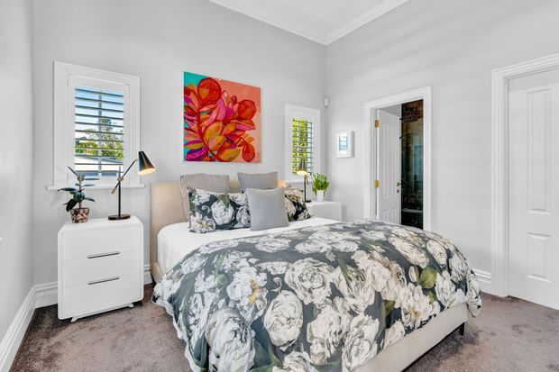 Bright, well-appointed bedroom with a large upholstered bed dressed in a dark floral duvet and layered cushions. The room has soft light-grey walls, white decorative trims and doors, carpeted flooring, plantation shutters, bedside storage, artwork, and a small desk lamp. An open doorway leads to an adjoining space or bathroom.