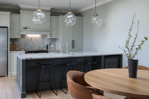 A refined kitchen and dining area with pale grey-green shaker cabinetry, a dark island with marble benchtop, integrated seating, and three spherical pendant lights. A round timber dining table with leather chairs occupies the foreground, while a potted branch adds a natural accent.
