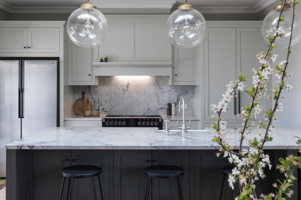 A refined contemporary kitchen with a large marble island, dark paneled cabinetry, pale grey perimeter joinery, integrated refrigeration, a range cooker, and a veined stone splashback. Three clear globe pendant lights hang above the island, with dark stools beneath; flowering branches partially frame the foreground.