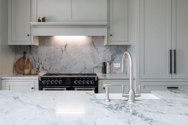 A refined traditional-style kitchen featuring pale grey shaker cabinetry, extensive white marble-look surfaces, a large island with an undermount sink and tall gooseneck faucet, a black range cooker, and a softly veined marble splashback illuminated beneath a mantel-style rangehood surround.