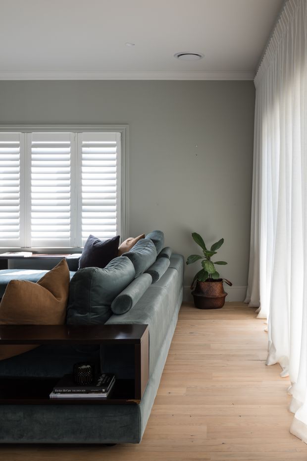 A refined living room with a large sectional sofa in muted blue-grey upholstery, layered neutral and tan cushions, and a dark timber console table. Pale timber flooring, grey-green walls, white plantation shutters, sheer full-height curtains, and a potted plant create a calm, softly lit interior. Decorative ceiling cornices and recessed air-conditioning are visible.