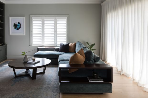 A refined living room with a deep blue-green sectional sofa, dark wood oval coffee table, and a dark wood console-style sofa table. Grey walls, white plantation shutters, full-height sheer curtains, pale timber flooring, built-in dark cabinetry, and a large neutral rug create a calm, softly lit interior.