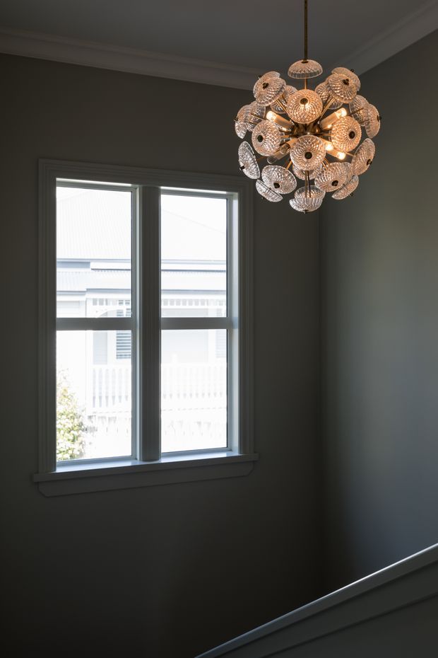 A dim, refined stairway or landing with dark grey painted walls, white decorative ceiling cornices, and a pair of tall white-trimmed windows. A sculptural clustered pendant light with numerous perforated metallic shades hangs near the upper right, while a white stair handrail runs diagonally across the lower frame. Bright neighboring architecture is visible through the windows.
