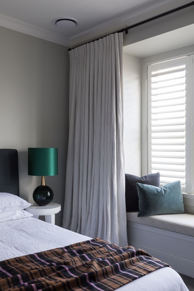 A refined bedroom interior with a partially visible bed, grey upholstered headboard, white bedding, and a dark patterned throw. Floor-to-ceiling light grey curtains frame a bright window with white plantation shutters and a built-in upholstered window seat. A small round white bedside table holds a dark glass lamp with a green cylindrical shade. The room has soft grey walls, white trim, and a calm, layered palette.