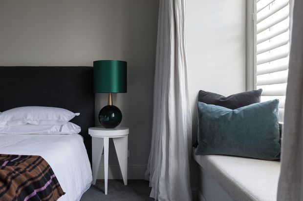 A compact, contemporary bedroom corner with a partially visible bed, dark upholstered headboard, white bedding, and a small white geometric bedside table. A dark green table lamp sits beside the bed, while a deep window seat with blue-grey cushions occupies the right side beneath white plantation shutters. Soft grey curtains and neutral finishes create a calm, refined atmosphere.
