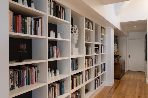 A bright residential hallway lined with extensive custom built-in white shelving filled with books and decorative objects. The timber floor, neutral walls, recessed ceiling lights, and distant entry door create a warm, refined library-like corridor.