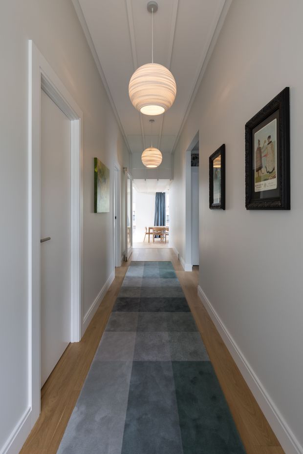 A bright, narrow residential hallway with white walls, detailed white ceiling moulding, timber flooring, and a long geometric runner rug in muted blue, grey, and green tones. White interior doors line the corridor, while framed artwork and mirrors decorate the right wall. Two warm spherical pendant lights lead toward a bright dining area at the far end.