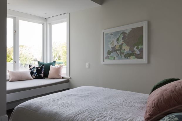 A calm, light-filled bedroom with a built-in window seat, pale upholstered bedding, and soft pastel cushions. Large corner windows bring in natural light, while a framed artwork hangs on the neutral wall. The room has a clean, contemporary appearance with minimal decoration.