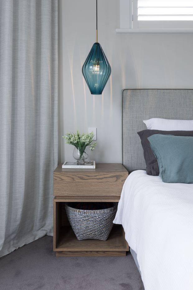 A calm, contemporary bedroom vignette featuring a grey upholstered bedhead, white bedding, blue-grey accent cushions, a warm timber bedside table with open shelving, and a distinctive teal glass pendant light. Floor-to-ceiling grey curtains, grey carpet, pale grey walls, and a partially visible shuttered window complete the scene.
