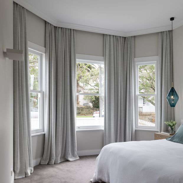 Bright, refined bedroom with three tall bay-style windows dressed in full-length light grey curtains and matching roller blinds. The room has pale walls with decorative crown moulding, soft grey carpet, a white bed with light blue-green cushions, a timber bedside table, and a sculptural teal pendant light. Mature greenery is visible outside the windows.
