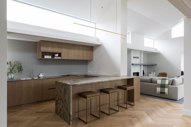 Open-plan contemporary kitchen with warm timber cabinetry, a large veined stone island with bar seating, and light herringbone timber flooring. The kitchen opens to a bright living area with high ceilings, clerestory windows, and a neutral sofa. Minimal pendant lighting and clean white walls create an airy, refined interior.