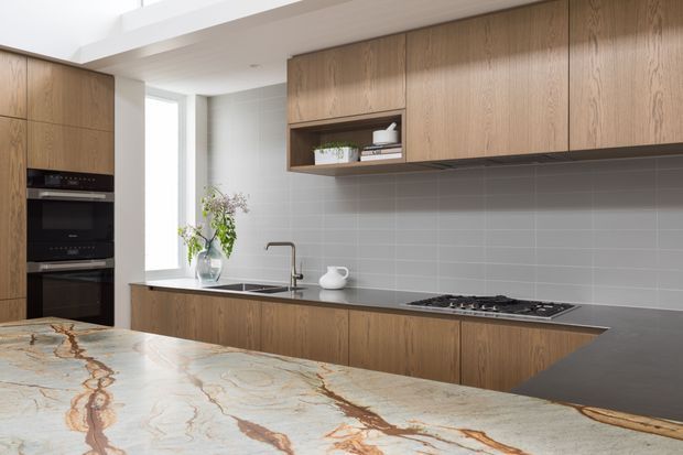 Contemporary kitchen with extensive warm wood cabinetry, integrated black appliances, a long grey tiled splashback, dark stone work surfaces, and a large island featuring dramatic veined stone. The space is bright and streamlined, with a skylight or high clerestory opening providing natural light.