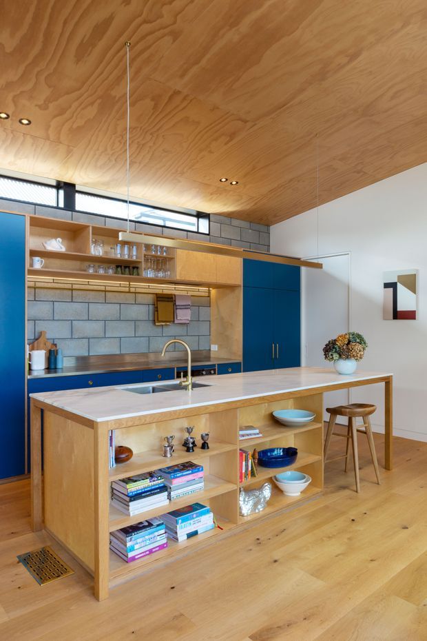 A compact, open-plan kitchen with a timber-lined ceiling, warm timber cabinetry and flooring, deep blue lower and tall cabinets, and a grey blockwork backsplash. The kitchen features a central island with a pale stone benchtop, integrated sink and brass-toned tap, open display shelving, pendant lighting, and two timber stools. High clerestory glazing brings in natural light, while books, crockery, and small decorative objects occupy the open shelves.