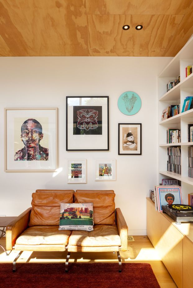 A compact, sunlit living room or reading space with a vintage cognac leather loveseat, framed contemporary artwork arranged on a white wall, extensive built-in white shelving filled with books and records, and a warm plywood-lined ceiling with recessed spotlights. A timber cabinet runs beneath the shelving, while a deep red rug and strong afternoon sunlight add warmth and contrast.