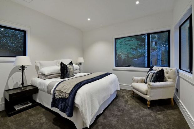 A bright, compact bedroom with a neatly made bed, white bedding, navy and grey-blue throws, bedside tables, table lamps, and a tufted armchair. Large black-framed windows overlook leafy greenery, while recessed ceiling lights and white walls create a clean, contemporary atmosphere.