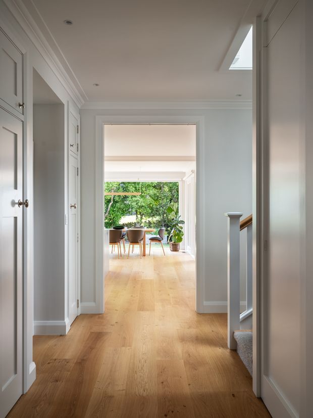 Bright, refined residential hallway with pale painted walls, detailed white trim and panelled cabinetry, warm light timber flooring, and a carpeted staircase with a white newel post. The corridor opens into a sunlit dining or living area with garden views and indoor plants. A rooflight adds natural light to the hallway.