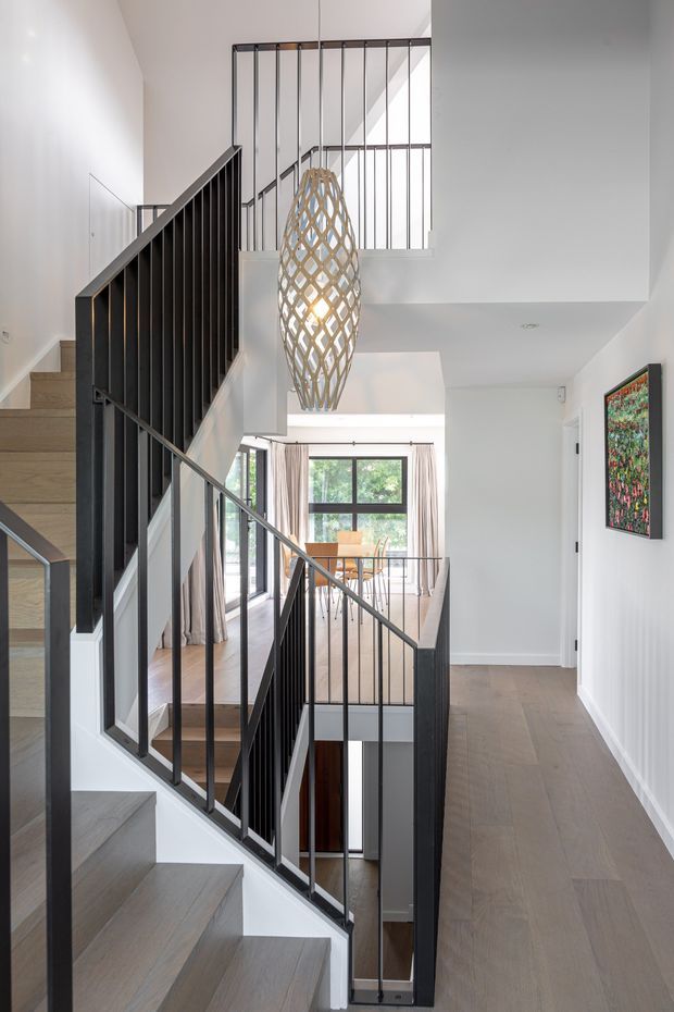 Bright contemporary multi-level stairway and landing with pale timber stair treads and flooring, crisp white walls, and slim black metal balustrades. A sculptural woven pendant hangs through the stair void, while large glazed doors and windows bring in natural light. A colourful framed artwork provides a small accent on the right wall.