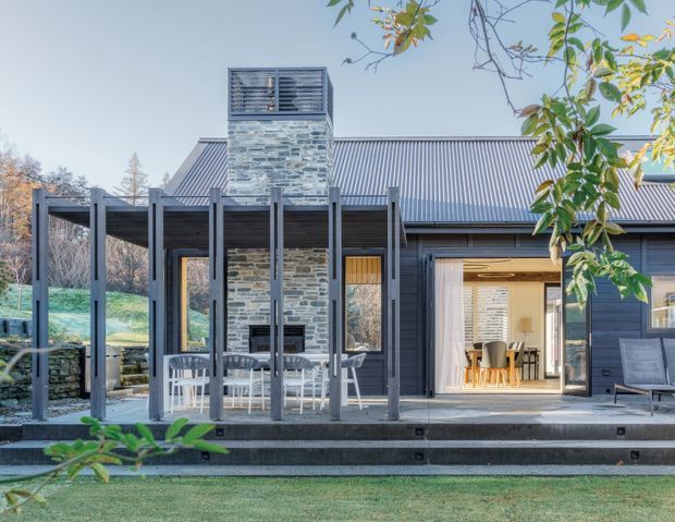 Exterior view of a contemporary single-storey home with dark vertical cladding, a corrugated metal pitched roof, and a prominent natural-stone chimney. A covered timber deck with slender posts extends across the facade, with outdoor dining and lounge areas visible. Large glazed openings reveal an interior dining space, while landscaped grounds and mature trees surround the house.