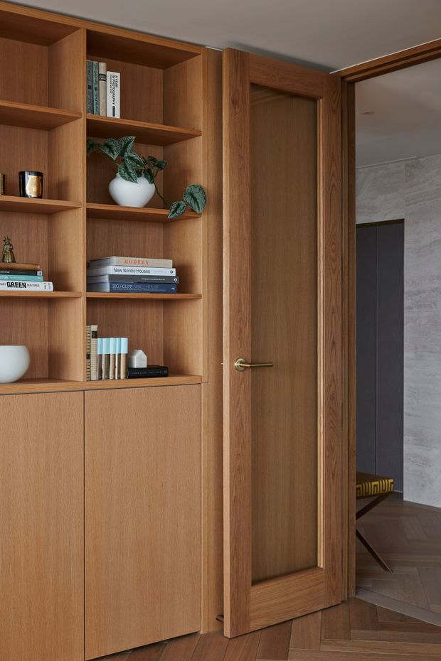 Interior view of a warm timber-lined built-in shelving and storage unit beside a framed timber door. Open shelves display books and small decorative objects, while the lower section has closed cabinetry. A glimpse through the doorway shows a darker adjoining room with patterned flooring and a textured light wall finish. The floor is laid in a dark timber herringbone pattern.