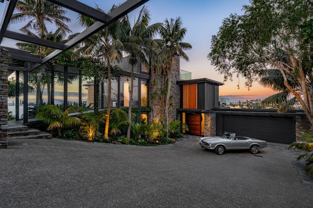 A modern tropical residential exterior at dusk, viewed from a paved driveway. The home features dark vertical cladding, extensive floor-to-ceiling glazing, a covered steel-framed outdoor structure, stone-clad columns, and a black garage door. Lush palms and layered planting surround the house, with a distant coastal or urban outlook visible beyond the property. A classic convertible is parked in the driveway.