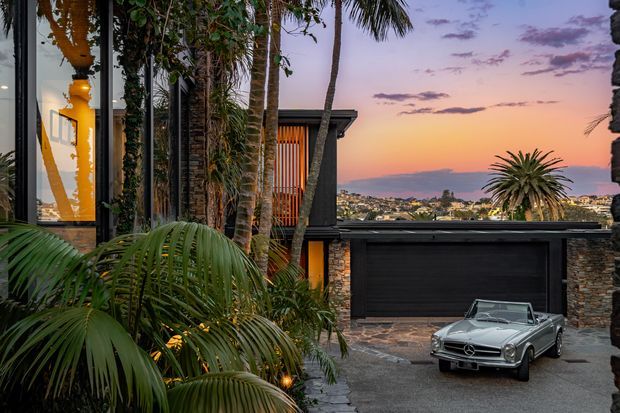 Twilight exterior view of a contemporary multi-level residence with dark cladding, stone walls, vertical timber screening, tropical planting, and a large integrated garage. A luxury car is parked in the driveway, while a coastal or hillside neighbourhood is visible beyond the property.