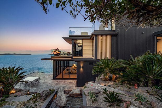 A modern multi-level coastal home set on a rocky landscaped waterfront site at dusk. The dark vertical-clad facade combines with warm timber screening, extensive glazing, cantilevered terraces, and transparent balustrades. Outdoor terraces and stepped stone paths connect the building to the sea, with subtropical planting throughout.