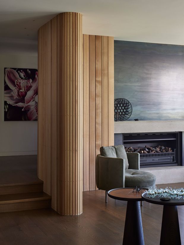 Refined contemporary living room featuring a curved vertical timber screen that divides the stairway and seating area. Dark timber flooring, an upholstered armchair, round side tables, a built-in fireplace, and a large atmospheric abstract wall artwork create a warm, sophisticated interior.