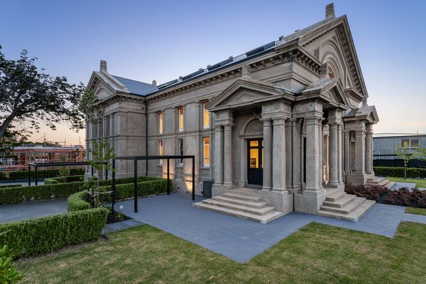 Exterior view of a formally composed, symmetrical stone-clad building with a prominent classical entrance portico, paired columns, triangular pediments, elaborate cornices, pilasters, and arched decorative detailing. The building sits within a manicured landscape with clipped hedges, paved paths, lawn, and contemporary surrounding structures. Warm interior lighting is visible through the windows and entry door at dusk.