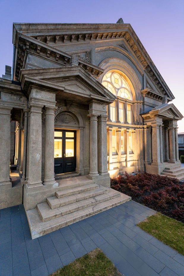 Exterior architectural photograph of an ornate stone building with monumental classical detailing. The facade features a large arched stained-glass window, Corinthian-style columns, layered pediments, heavy cornices, decorative dentils and carved reliefs. Broad stone steps lead to dark double doors, with paved paths and low landscaping in the foreground. The image is captured at dusk, with warm light glowing through the windows.