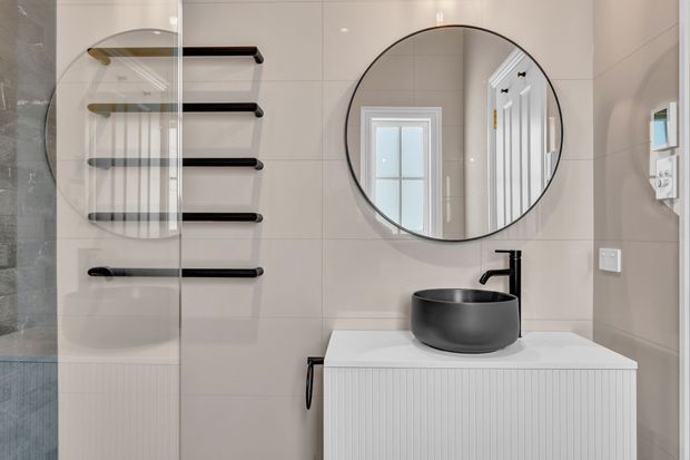 Contemporary bathroom featuring large pale wall tiles, a frameless glass shower enclosure, black heated towel rail, round black-framed mirror, matte black vessel basin and tap, and a white vertically fluted vanity.