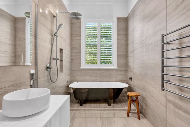 Bright, refined bathroom featuring large beige stone-look wall and floor tiles, a freestanding dark-sided bathtub beneath a shuttered window, a white vessel basin on a built-in vanity, a glass-enclosed shower, and a chrome heated towel rail. The space has a clean, contemporary-traditional feel with natural light and neutral finishes.