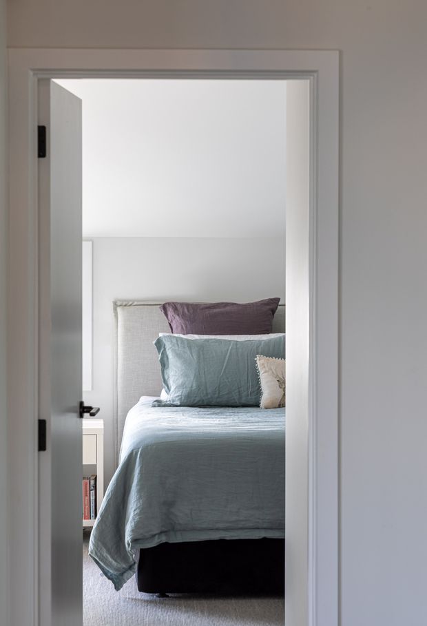 View through an open white-trimmed doorway into a compact, softly lit bedroom. The room contains a neatly made bed with pale blue-grey linen bedding, layered blue, mauve and cream pillows, a dark upholstered bed base, light carpet and a small white bedside unit. The setting has simple white walls and restrained contemporary styling.
