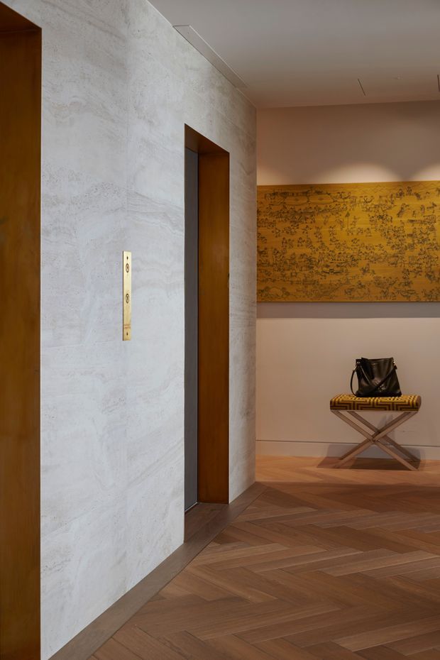 Warmly lit interior hallway or entry area featuring a pale textured stone or plaster wall, tall dark timber door openings, and a rich herringbone timber floor. A brass-toned vertical wall control or lift panel is visible, while the adjoining wall displays a large ochre decorative artwork above a folding stool with a black bag. The composition emphasizes refined material detailing and a contemporary, gallery-like atmosphere.