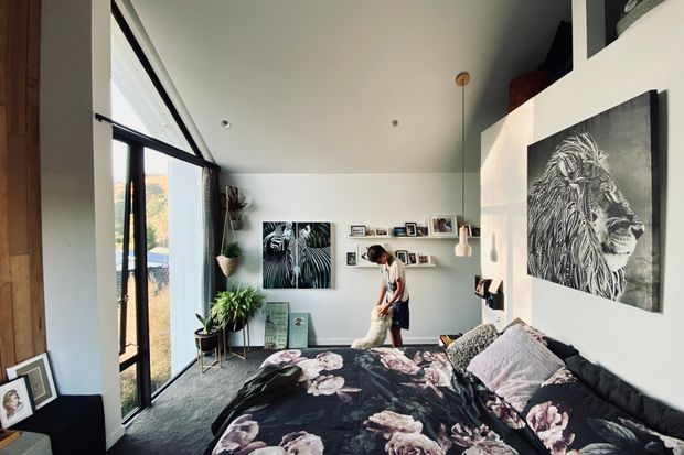 A bright contemporary bedroom with a large bed in the foreground, floral dark bedding, abundant indoor plants, framed artwork, and a narrow picture ledge. Floor-to-ceiling black-framed glazing runs along the left side, bringing in natural light and views of greenery. The room has a high ceiling, pale walls, dark carpet, and a large monochrome lion artwork as a focal point.