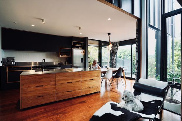 A contemporary open-plan kitchen and dining area with a large timber island, dark cabinetry, stainless-steel appliances, and expansive black-framed floor-to-ceiling glazing. Warm timber flooring continues into the living area, where a black leather lounge chair with a cowhide throw sits beside the windows. A dining table and chairs are visible beyond the island, with a double-height upper-level opening adding volume.