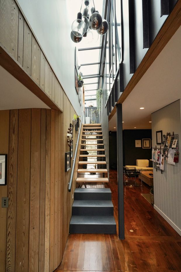 A narrow, double-height interior hall centred on an open-riser staircase. Warm timber wall panelling and polished timber flooring contrast with dark-painted stair treads, black structural framing, and a glazed roof/skylight that brings in abundant daylight. A sculptural cluster pendant hangs above the stair, while a living area and wall-mounted display board are visible to the right.