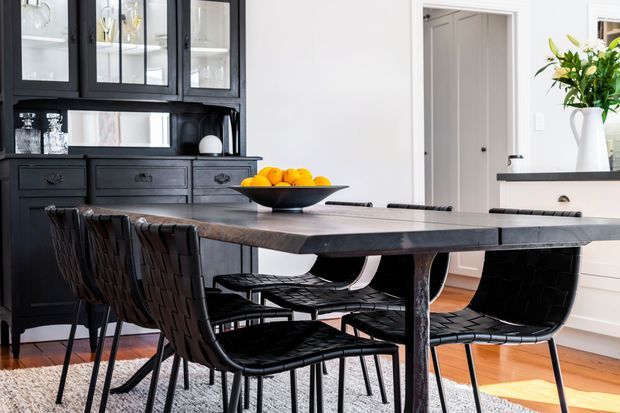 A refined dining area with a substantial dark timber dining table and woven black leather chairs. A black built-in cabinet with glass-front upper storage and drawers lines the left wall, while white cabinetry and a vase of greenery sit to the right. Warm timber flooring, a grey rug, and bright white walls create a high-contrast, elegant interior.