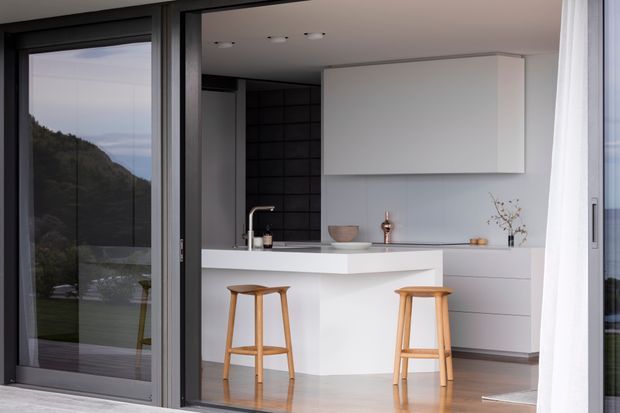 A contemporary minimalist kitchen viewed through large dark-framed glazing. The space features a white island with a slim benchtop, integrated white cabinetry, pale blue-grey walls, a dark tiled splashback, timber bar stools, light timber flooring, and sheer curtains. A mountain landscape is visible through the glazing.