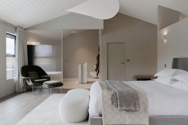 A refined contemporary bedroom with a low-profile upholstered bed, layered white bedding and textured throws, a rounded upholstered ottoman, and a black lounge chair with footstool. The room features pale timber flooring, soft neutral walls, a white slatted ceiling, full-height curtains, minimalist built-in elements, and sculptural ceiling lighting. A partially screened adjoining area is visible beyond the bedroom.