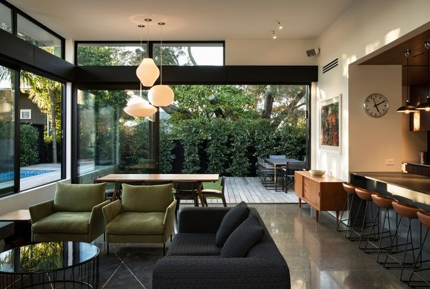 A bright, open-plan contemporary living and dining area with a connected kitchen and bar. Floor-to-ceiling black-framed glazing opens toward a timber deck and lush garden, with a glimpse of a swimming pool outside. The interior features a dark sectional sofa, olive-green lounge chairs, a timber dining table, a timber sideboard, polished dark flooring, pendant lights, and a kitchen island with bar seating.