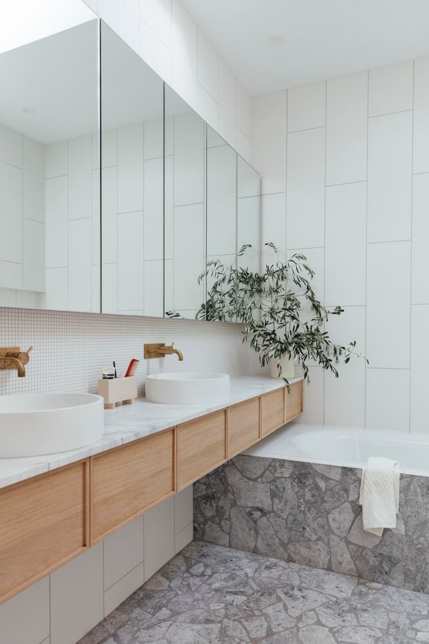 Bright contemporary bathroom with a double vanity, wall-to-wall mirrors, white geometric wall tiling, pale timber floating cabinetry, white vessel basins, brass wall-mounted taps, and a freestanding bathtub. The bath surround and floor feature irregular grey natural stone, while a leafy plant adds an organic accent.