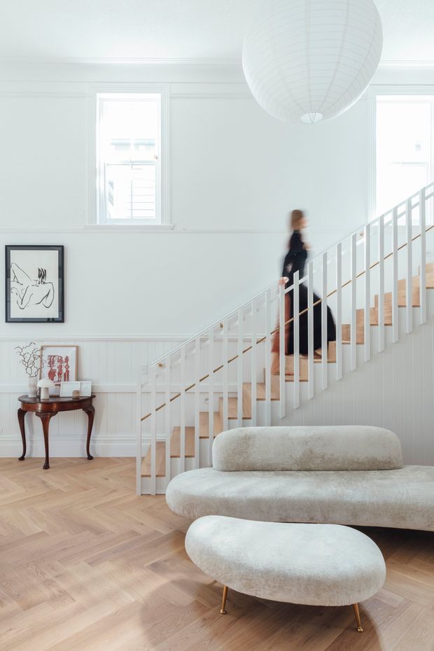 Bright, airy interior featuring a white-painted stairway with vertical balustrades, pale timber treads, white wall panelling and trim, and a large spherical pendant light. A curved cream boucle sofa and matching ottoman sit on a light herringbone timber floor, with a small dark timber console and framed artwork beside the stairs. The image presents an elegant open living and circulation space with a person ascending the staircase.