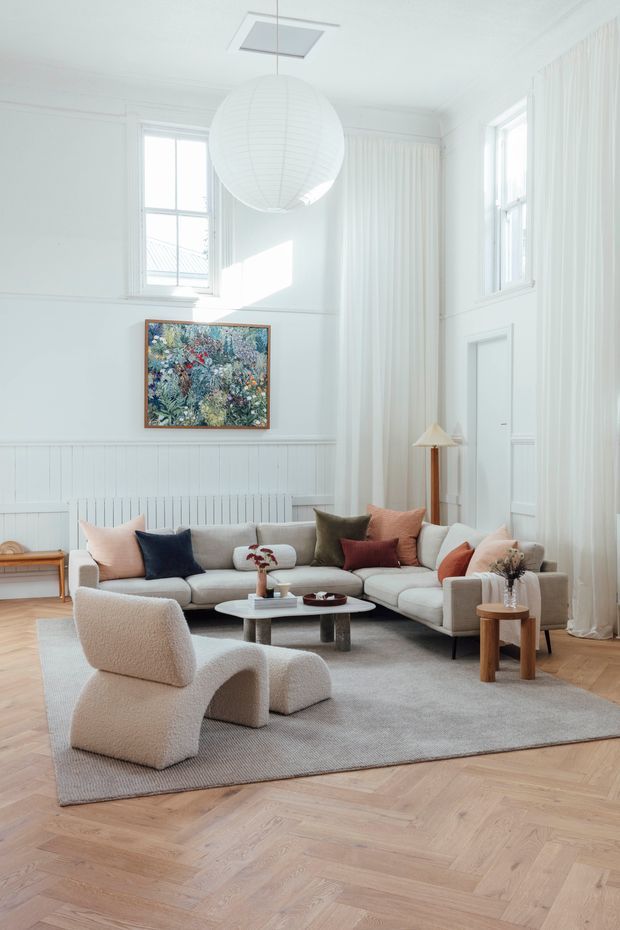 Bright, airy living room in a tall traditional-style interior, featuring white vertical wall panelling, decorative mouldings, large windows with sheer floor-to-ceiling curtains, and a white paneled door. The room is furnished with a cream sectional sofa, sculptural cream lounge chair, round coffee table, side tables, layered neutral cushions, a grey rug, and a large white globe pendant. Light timber herringbone flooring and a colourful botanical artwork add warmth and contrast.