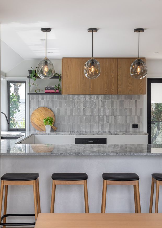A bright contemporary kitchen viewed from the dining area, featuring a long island with a pale stone benchtop and integrated seating. The kitchen has warm timber overhead cabinetry, a textured grey tiled splashback, pale grey cabinetry, black-framed glazing and doors, and three large smoked-glass pendant lights. Timber bar stools and the edge of a dining table are visible in the foreground.