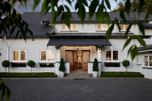 Front exterior view of a refined two-storey residence with a symmetrical pale stucco facade, dark pitched metal roofing, a projecting covered entry, timber double doors, and regularly spaced windows. Formal clipped shrubs and topiary flank the entrance, while foreground foliage partially frames the building.