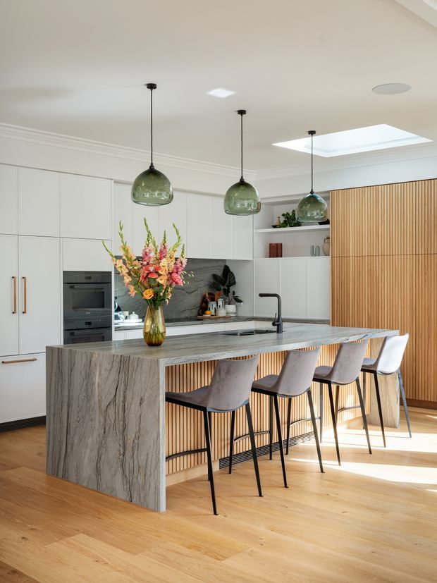 Bright contemporary kitchen with a large stone island and integrated breakfast bar, white handleless cabinetry, dark stone splashback, timber flooring, vertical timber slat detailing, and three green glass pendant lights. A skylight brings additional natural light into the space.