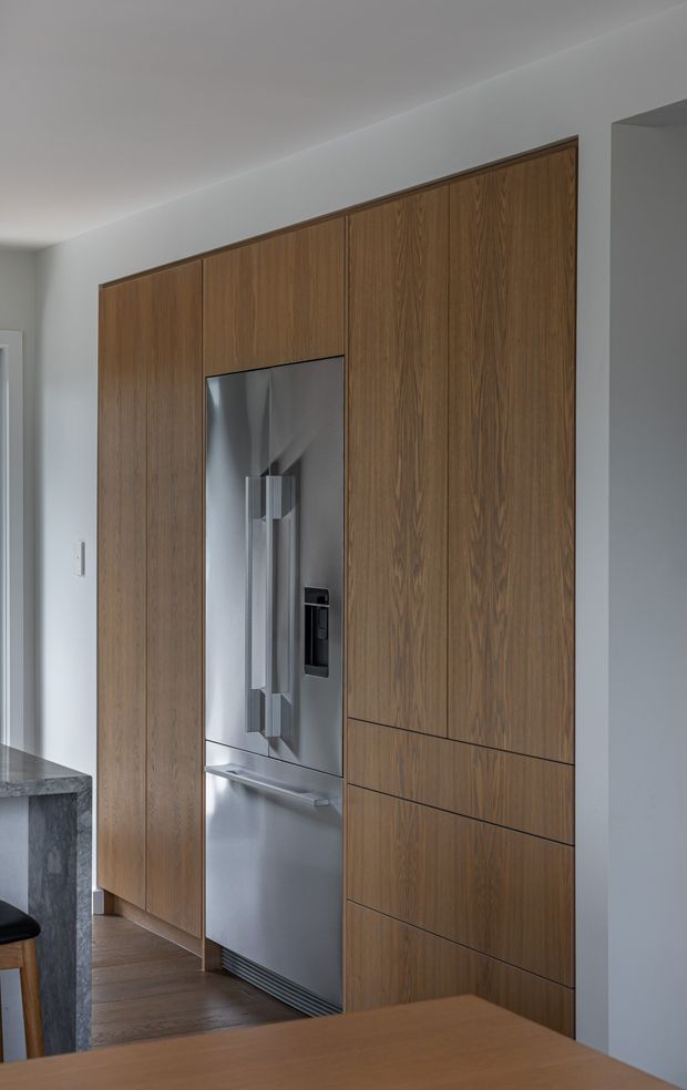 A close-up interior view of a modern kitchen wall featuring full-height dark timber-grain cabinetry with integrated drawers and a built-in stainless-steel refrigerator. A stone benchtop is partially visible at left, with timber flooring and white plaster walls and ceiling. The cabinetry is clean-lined and handleless, creating a streamlined contemporary fit-out.